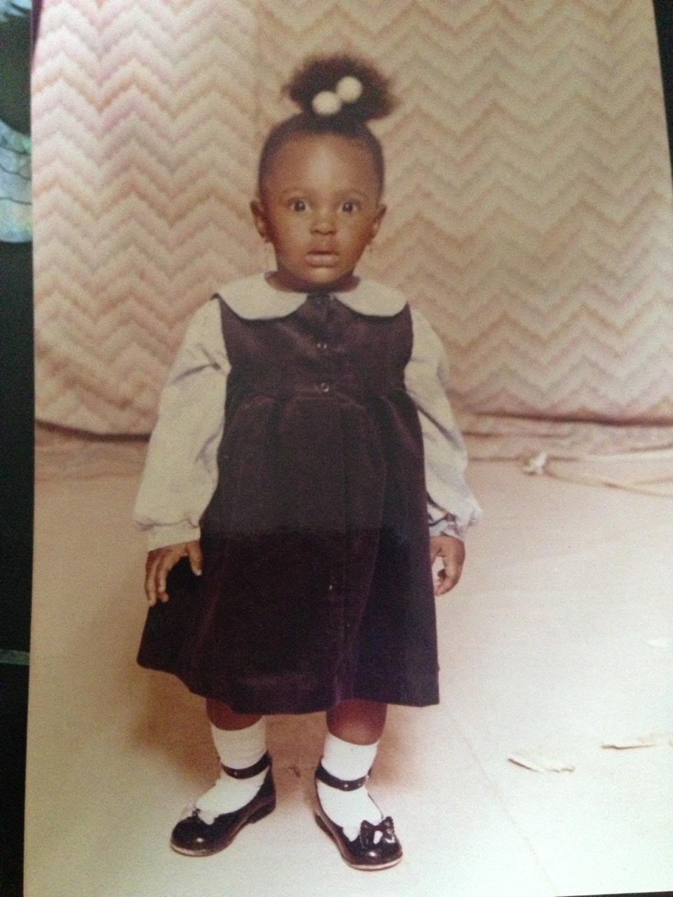 A toddler portrait of Amal in a dark dress and bow shoes